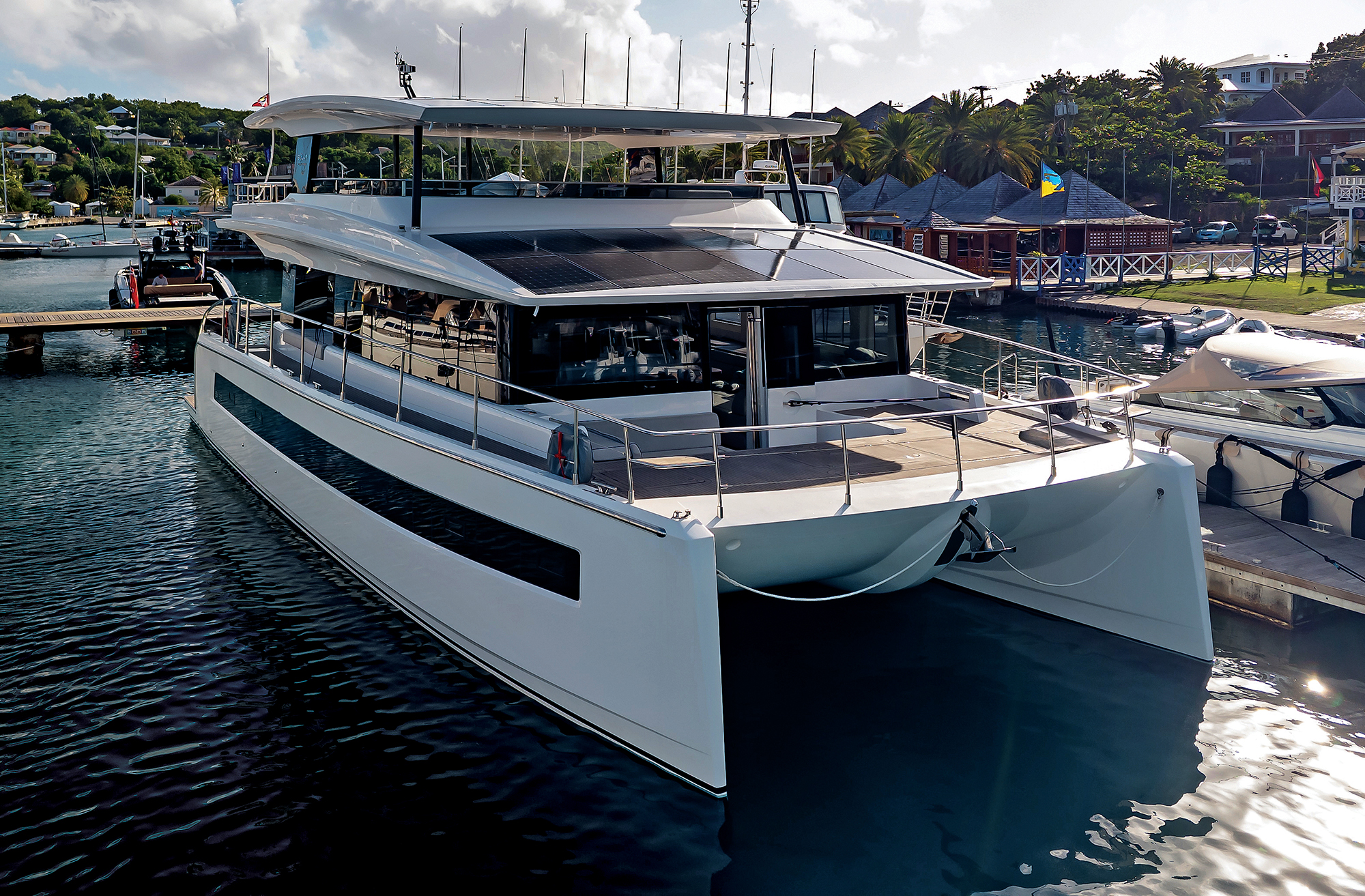A front-angle shot of a white Silent Yachts solar-electric catamaran docked at a marina, showcasing its wide multihull bow and solar panel array stretching across the upper decks.