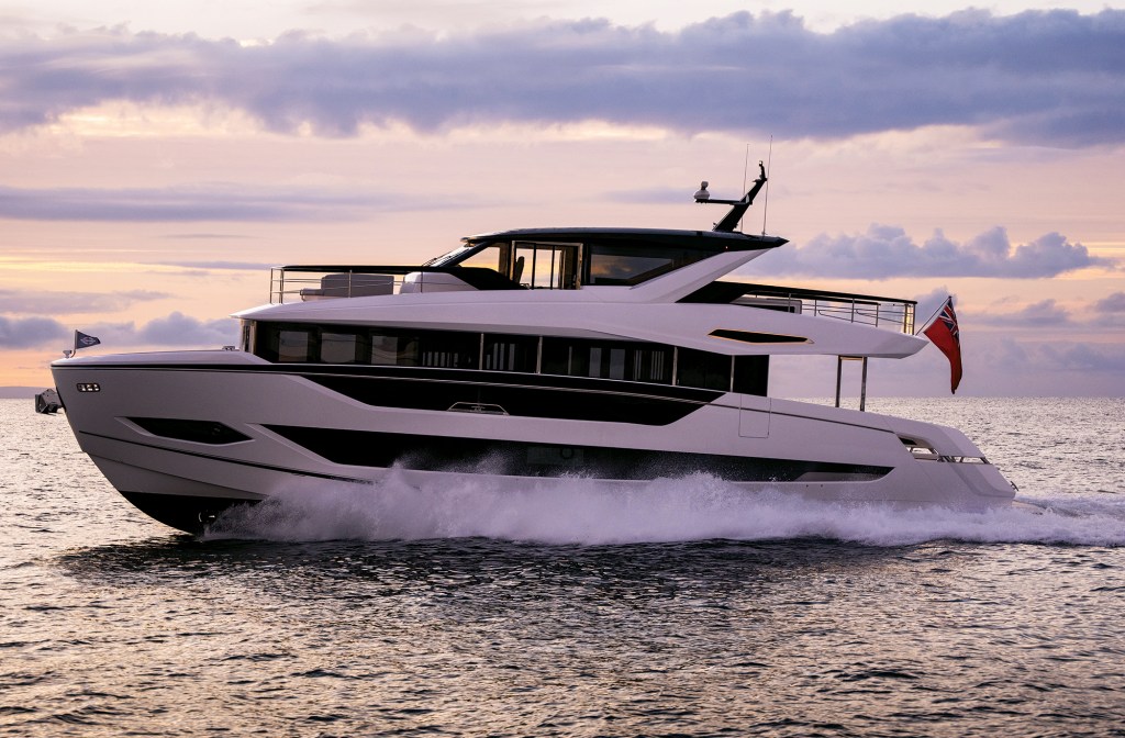 A side profile shot of a modern, white Sunseeker 82 Ocean luxury motor yacht cruising at speed across open water. The vessel features striking black-tinted window bands along its multi-deck hull and a black hardtop structure over its upper deck flybridge. It kicks up a substantial white spray and wake as it glides under a soft pink, purple, and gold sunset sky, with a British Red Ensign flag flying from the stern.