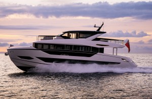 A side profile shot of a modern, white Sunseeker 82 Ocean luxury motor yacht cruising at speed across open water. The vessel features striking black-tinted window bands along its multi-deck hull and a black hardtop structure over its upper deck flybridge. It kicks up a substantial white spray and wake as it glides under a soft pink, purple, and gold sunset sky, with a British Red Ensign flag flying from the stern.