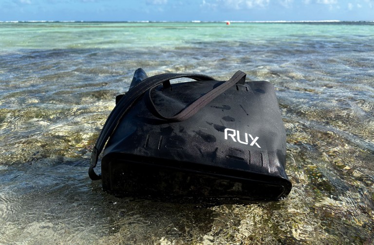 feat_duffel drybags A close-up, eye-level shot of a durable black RUX waterproof bag resting on a sandy bottom in shallow, crystal-clear ocean water. The bag features a minimalist design with white "RUX" lettering and a long black carry strap draped over the top. The translucent turquoise water ripples gently around the semi-submerged gear bag, with a calm sea horizon and a clear blue sky with light clouds visible in the background.