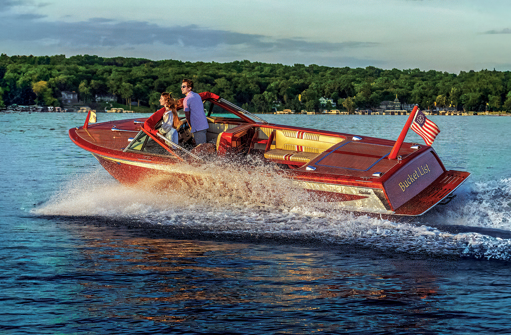 A classic mahogany wooden runabout boat named "Bucket List" cruising across a lake at dusk, throwing up a spray of white water.