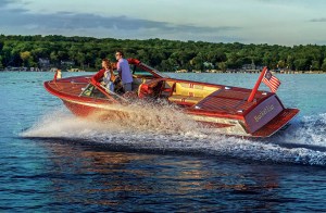 A classic mahogany wooden runabout boat named "Bucket List" cruising across a lake at dusk, throwing up a spray of white water.