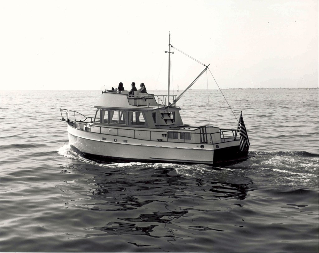 A vintage black-and-white photograph showing a classic Grand Banks 36 trawler underway on calm seas, viewed from the aft quarter with passengers visible on the flybridge and an American flag flying from the stern.