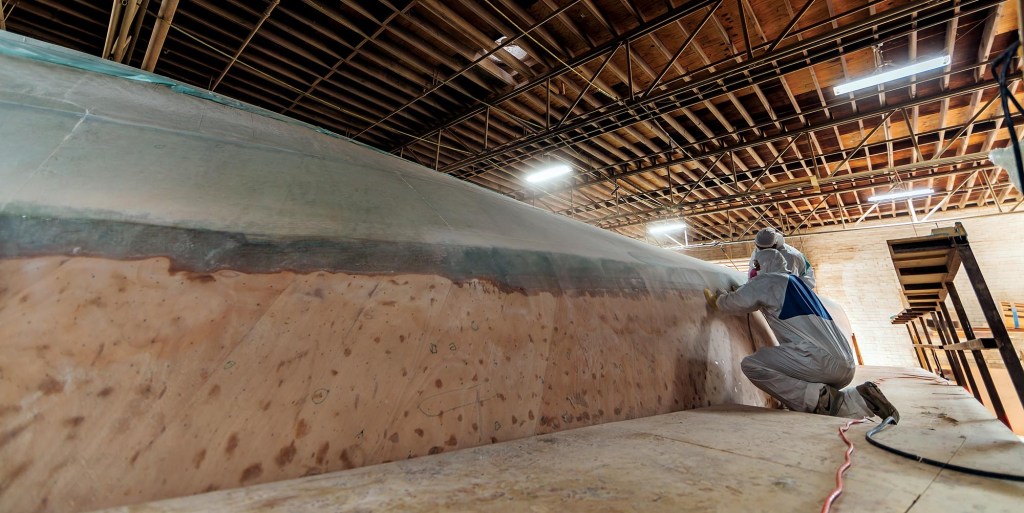 Shipyard workers wearing protective suits and masks working on the inverted wooden hull of a custom Jarrett Bay sportfishing boat inside a large boatbuilding facility.