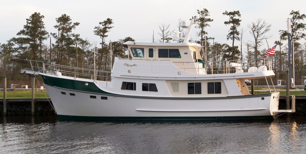 A side profile photograph of the Kadey-Krogen 55 Expedition yacht Olympia docked along a bulkhead with a pine forest shoreline in the background and an American flag flying from the stern.