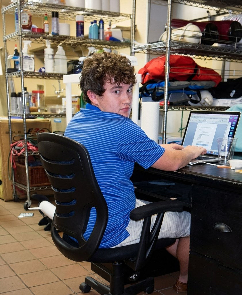 A young man sitting at a desk with an open laptop in an office or storage room lined with metal shelving, turning over his shoulder to look back at the camera.