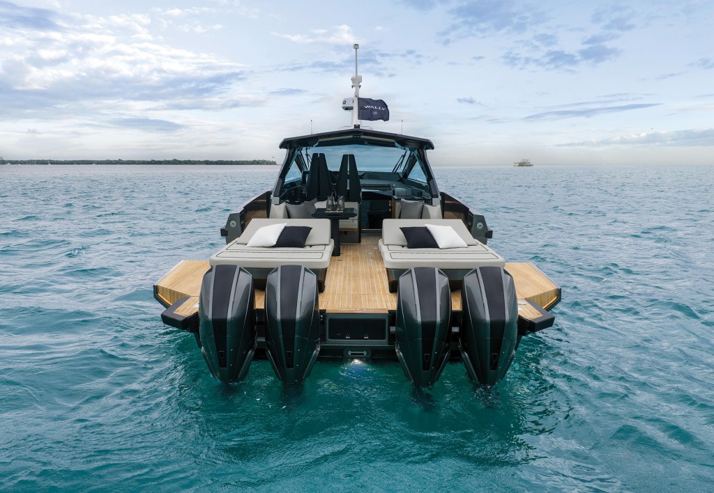 A stern-on view of the Wallypower 50X floating at rest in calm waters, highlighting its quad Mercury outboard engines, expanded fold-down teak side terraces, and twin sunpads leading toward the covered helm.