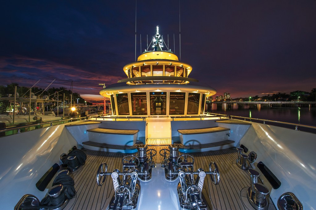 The teak foredeck and illuminated superstructure of the explorer yacht Beothuk docked at dusk, featuring dual chrome windlasses and ground tackle.