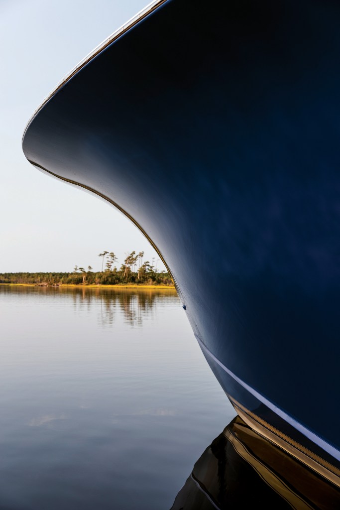 A close-up view of the dramatically flared bow of a dark-hulled Jarrett Bay custom sportfishing boat resting on calm water with a treed shoreline in the background.