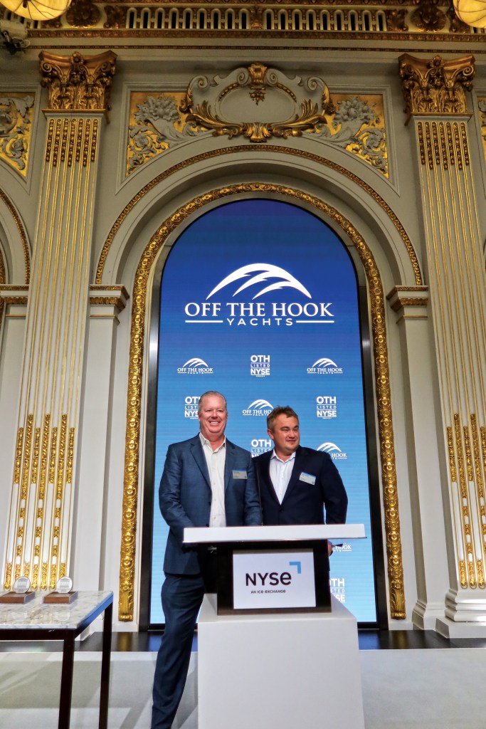 Two men in suits standing behind a podium bearing the New York Stock Exchange (NYSE) logo, celebrating Off the Hook Yacht Sales going public in an ornate hall with gold trim and a large blue digital backdrop displaying the company branding.