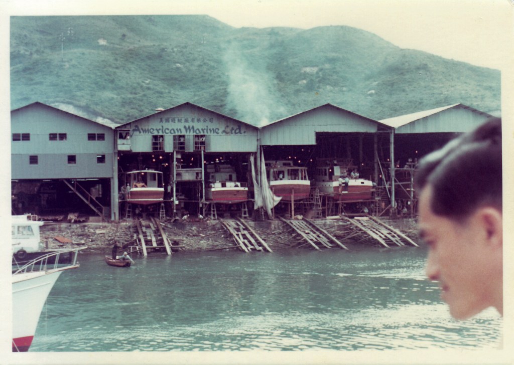 An archival color photograph showing the American Marine Ltd. shipyard facility in Hong Kong during the 1960s, with several wooden boats under construction in open-air covered bays along the waterfront and rolling green hills in the background.