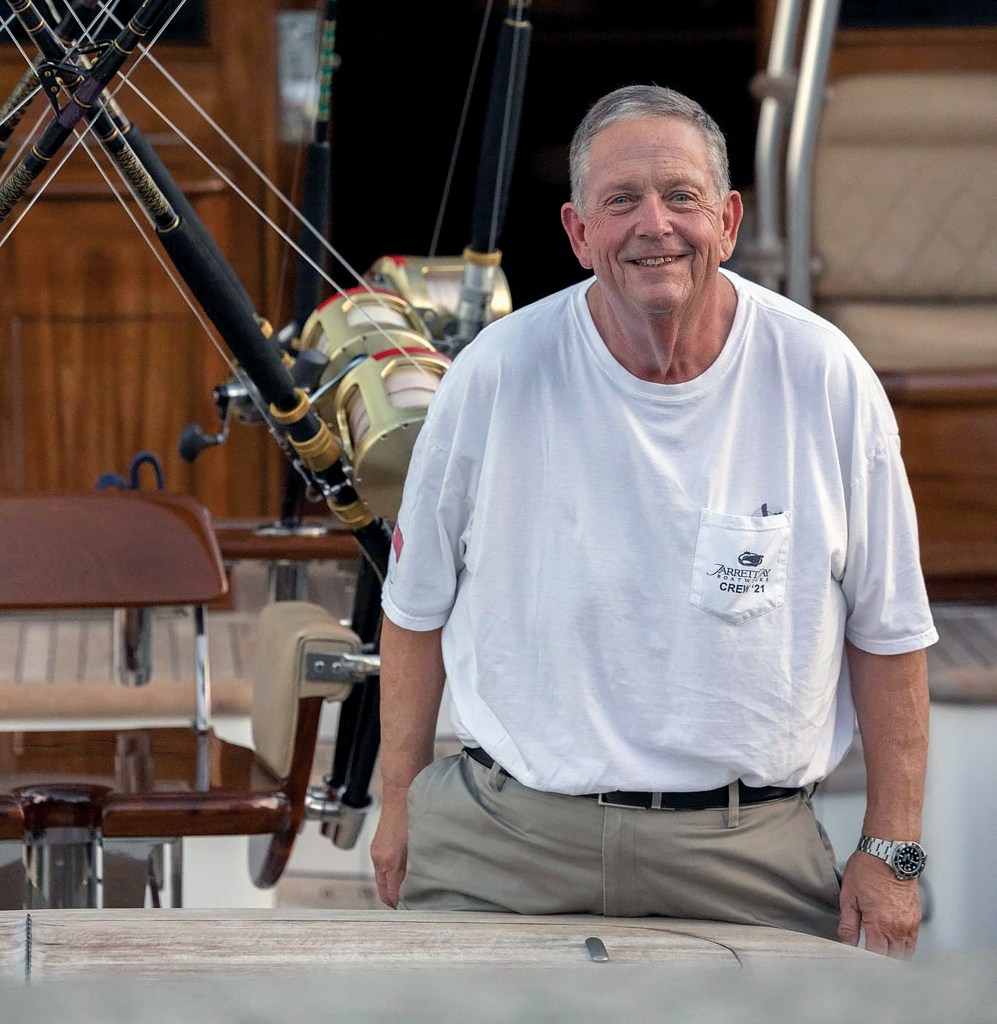 A smiling portrait of Jarrett Bay founder Randy Ramsey wearing a branded t-shirt aboard the teak cockpit of a sportfishing yacht, flanked by offshore fishing rods and reels.