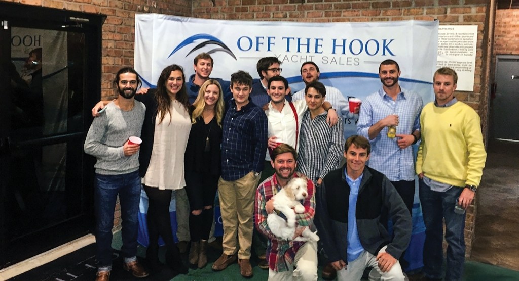 A group photo of the Off the Hook Yacht Sales team smiling together in front of a branded backdrop inside a venue with brick walls, including a man kneeling in front holding a fluffy white puppy.