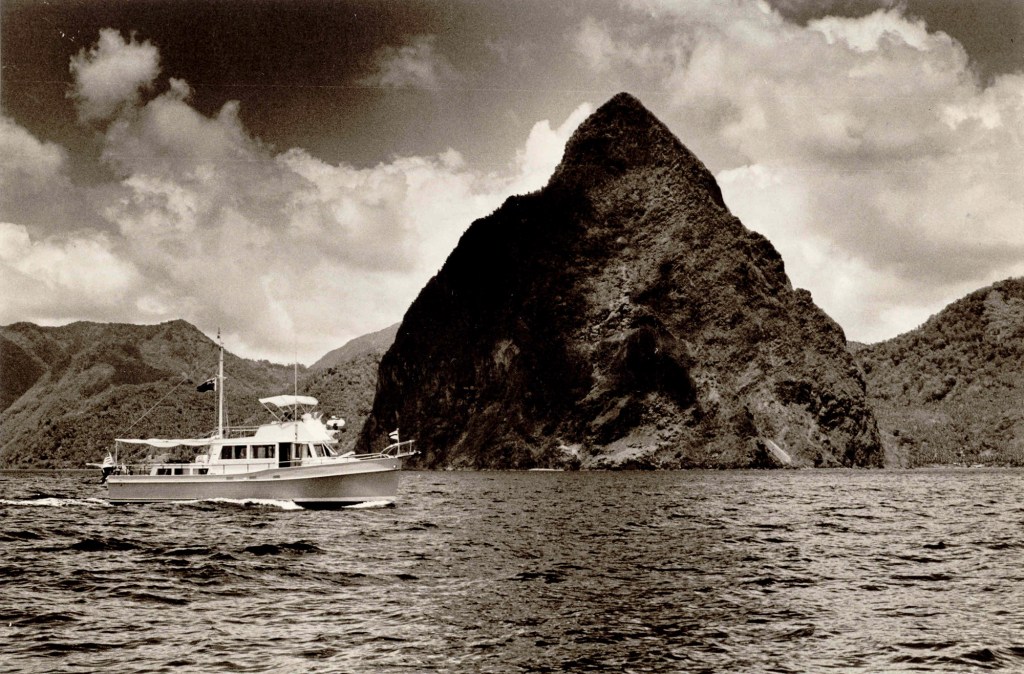 A vintage black-and-white photograph showing a classic Grand Banks trawler cruising past Petit Piton in Saint Lucia under a cloudy sky.