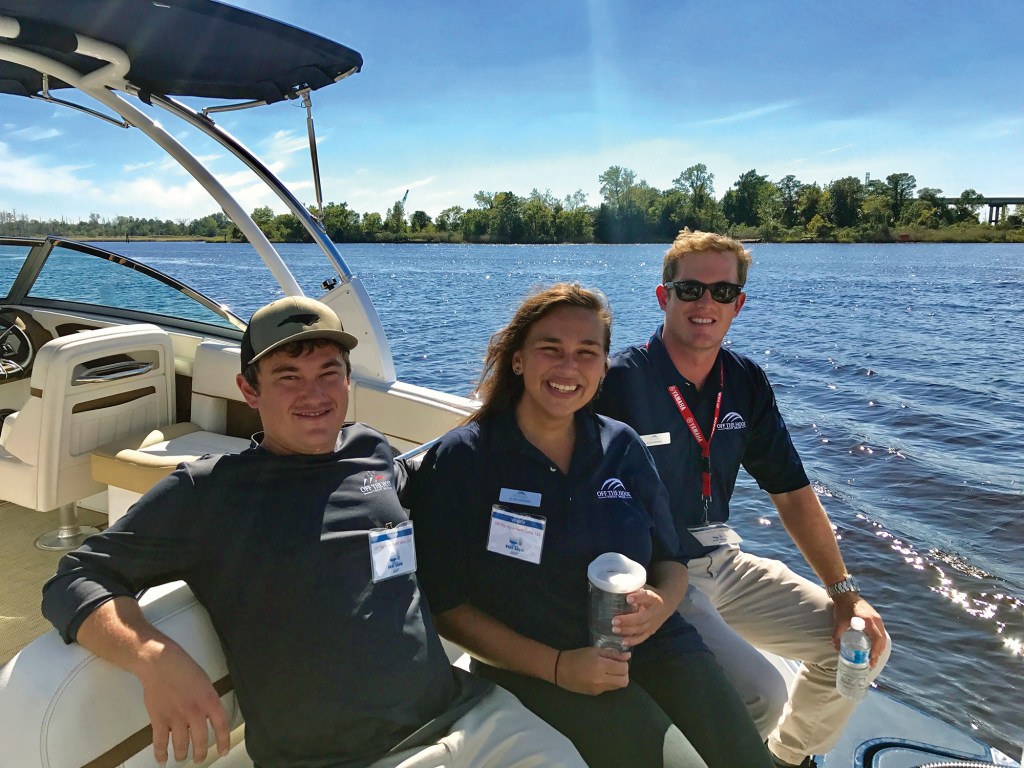 Three young adults wearing Off the Hook Yacht Sales shirts and event name badges sitting together on the stern seating of a powerboat under a bright, clear blue sky.