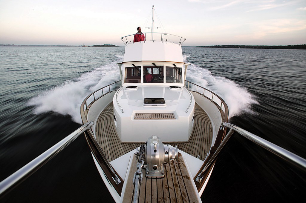 A bow-facing wide-angle shot of a Grand Banks motor yacht cutting through water, showing its teak foredeck, anchor windlass, superstructure, and captain standing at the flybridge helm under a soft evening sky.