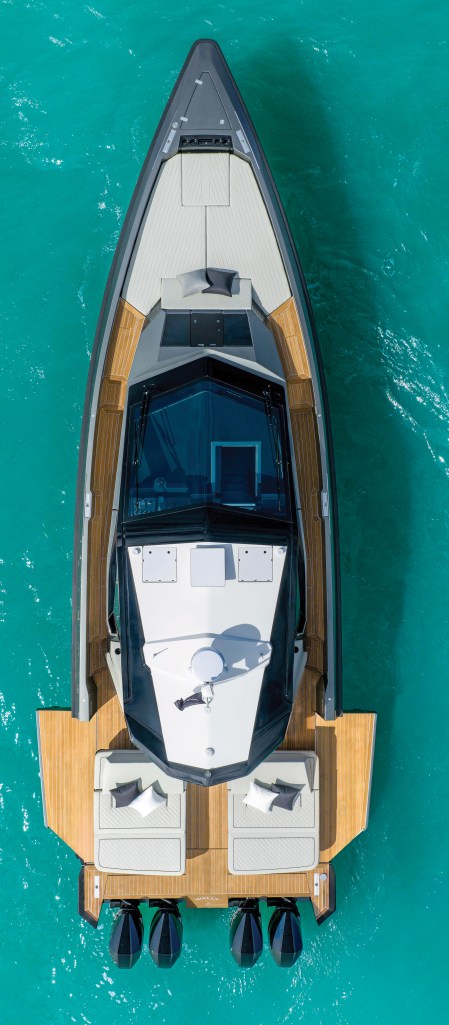 A top-down aerial view of the Wallypower 50X anchored in clear turquoise water, showcasing its sharp plumb bow, spacious teak side decks, fold-down aft terraces, and quad Mercury outboard configuration.