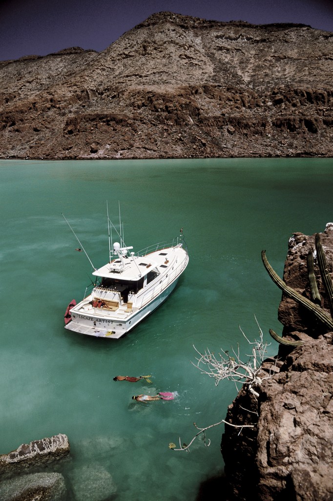 An aerial high-angle view showing an Eastbay 49 yacht named Escape Artist anchored in clear, turquoise water near a rocky desert cove, with two snorkelers swimming nearby.