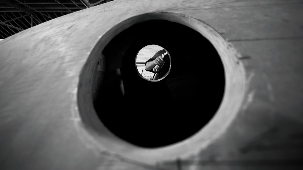 A black-and-white perspective shot through a circular cut-out on a wooden boat hull in construction, showing a craftsman holding a tool on the other side inside the boatyard.