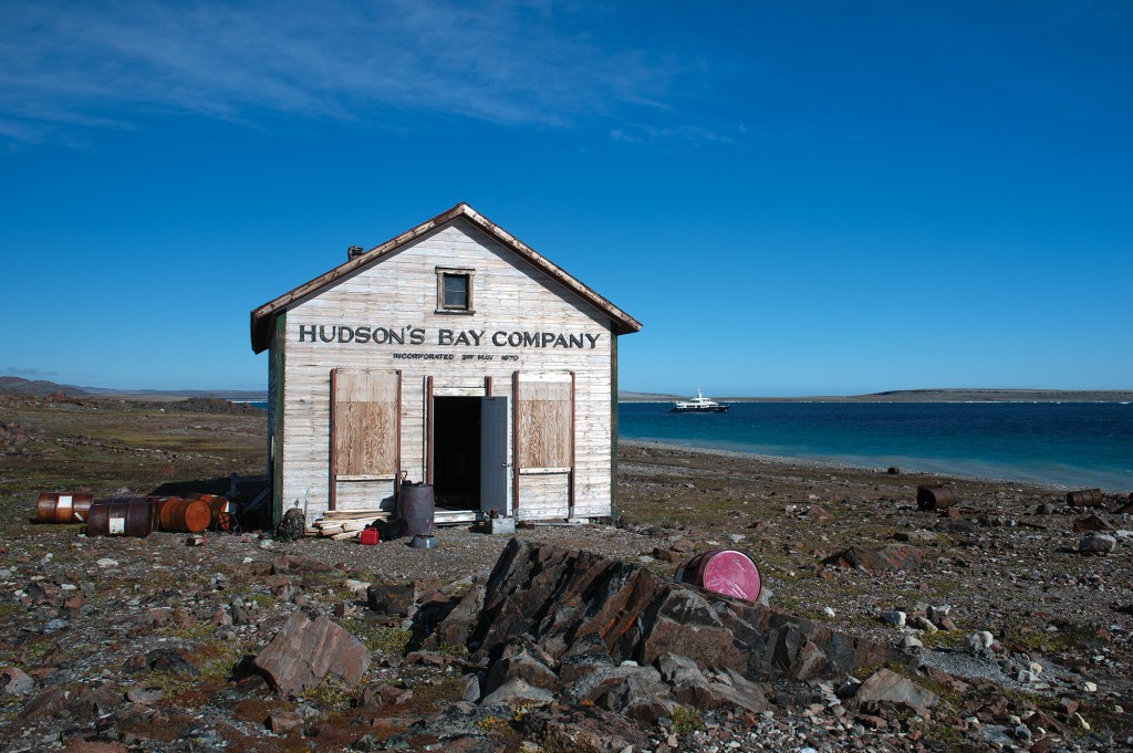 A historic, wooden Hudson's Bay Company trading post on a rocky Arctic shore with the explorer yacht Beothuk anchored in the waters offshore.
