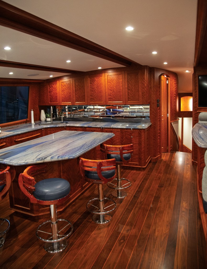 The custom galley interior of a Jarrett Bay yacht featuring polished blue marble counter surfaces, rich wooden cabinetry, bar stools with leather seats, and hardwood flooring.