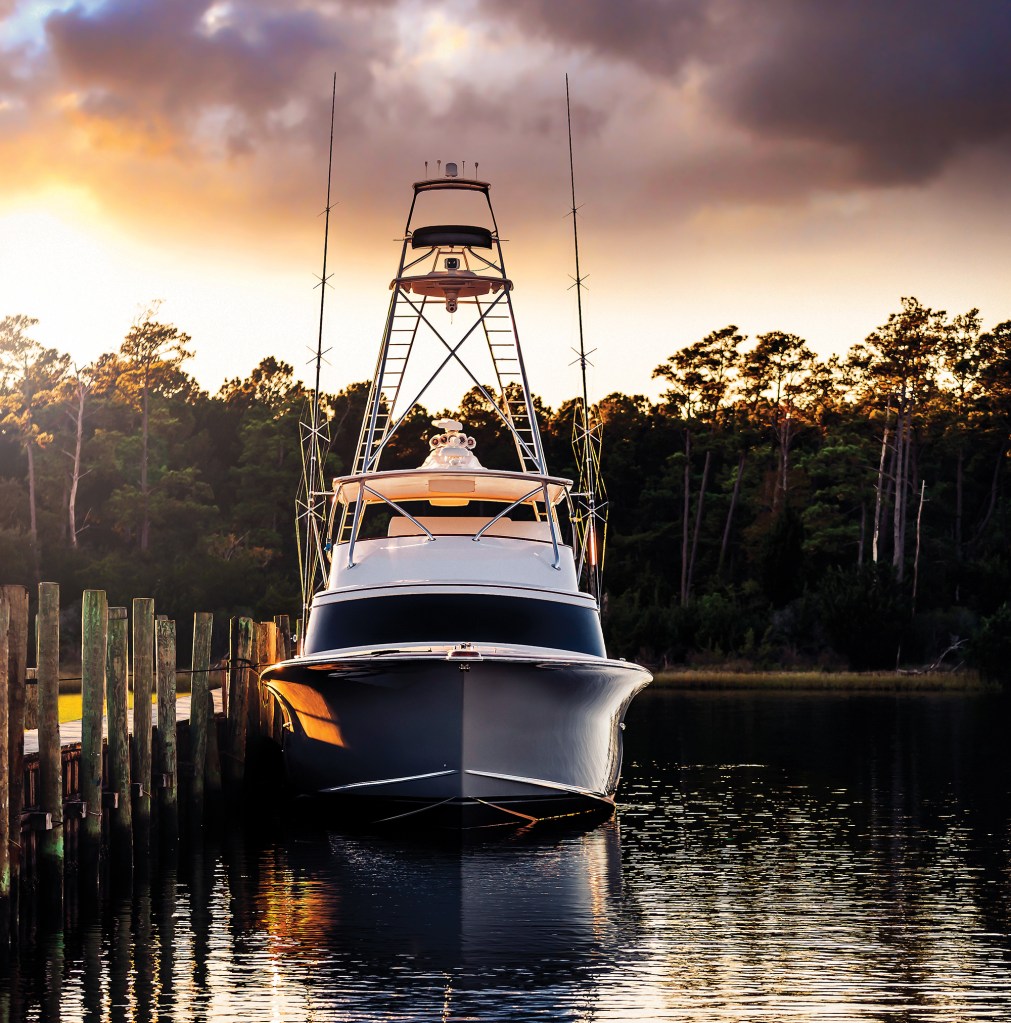 A Jarrett Bay sportfishing boat with a tall tuna tower docked beside a wooden pier at sunset, reflecting warm light across calm water in front of a tree-lined shoreline.
