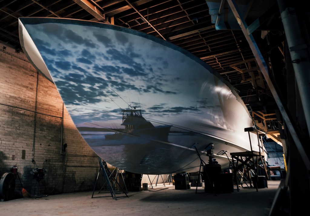 A dramatic view of a custom boat hull inside a shop, illuminated by a projected image of a sportfishing yacht underway at sunset across its smooth white surface.