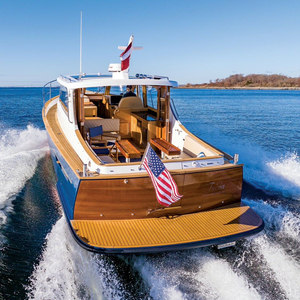 A stern view of a Duchy 35 motor yacht underway, highlighting its wooden transom with an American flag, teak swim platform, open cockpit seating, and teak side decks.