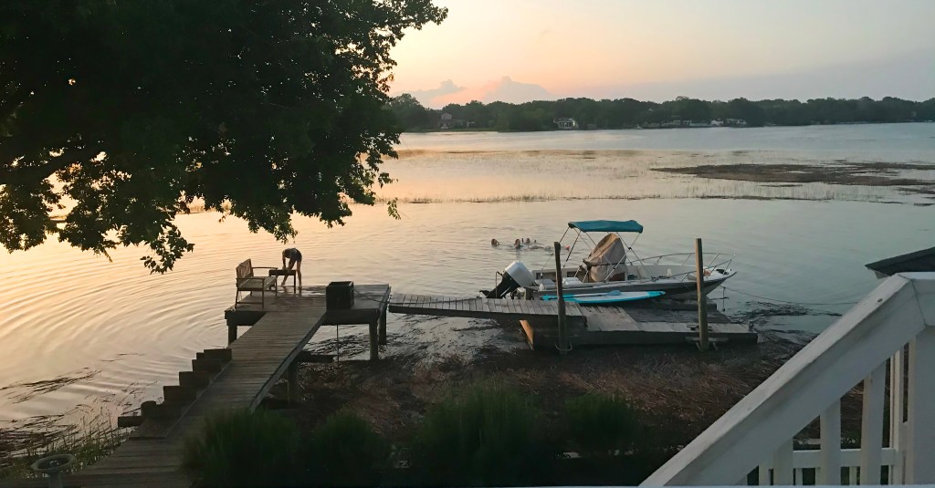 A wooden fixed dock leading to a floating dock structure at sunset, with a small skiff moored alongside, people swimming in the calm water nearby, and trees along the shore.