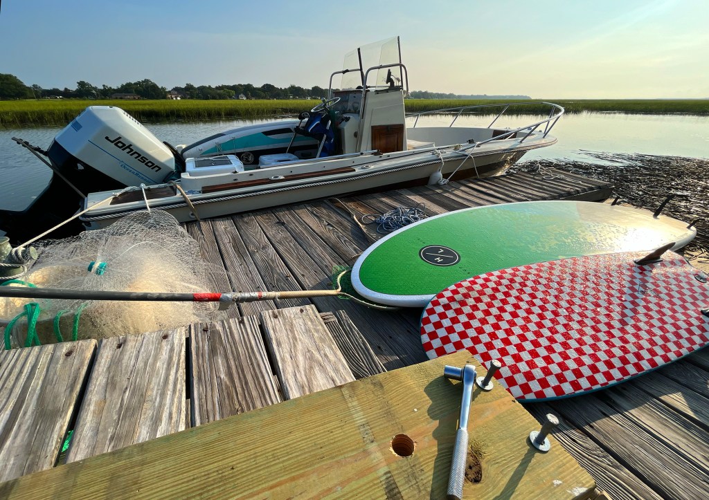 Tools, lumber, paddleboards, and a cast net laying on a wooden floating dock next to a center-console skiff tied up along a tidal marsh.