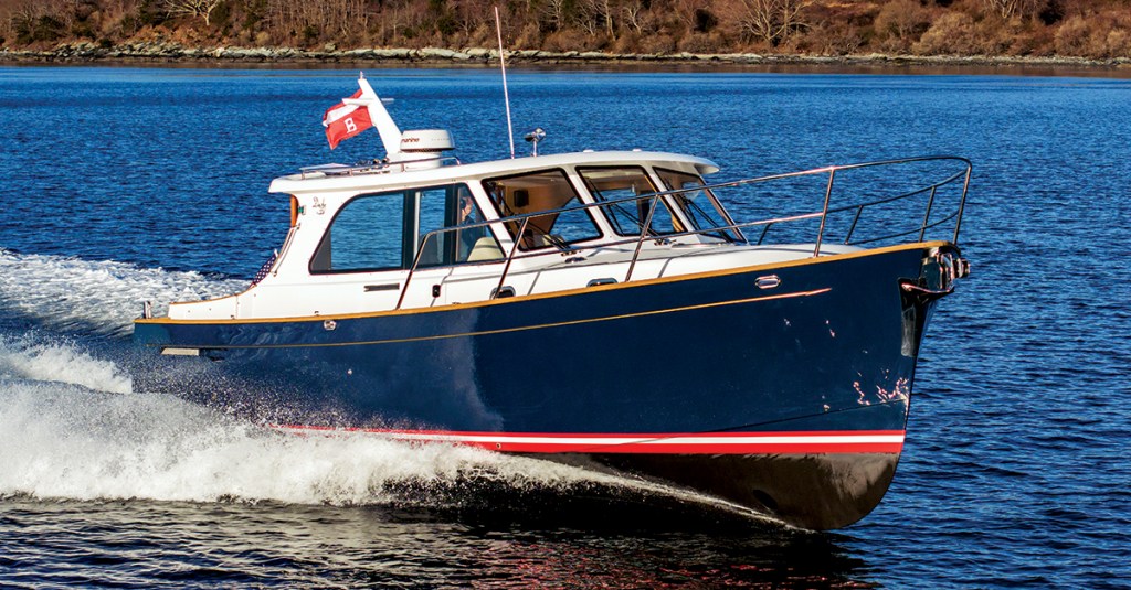 A navy blue and white Duchy 35 motor yacht with a red boot stripe cruising at speed through open blue water, leaving a white wake with a wooded shoreline in the background.