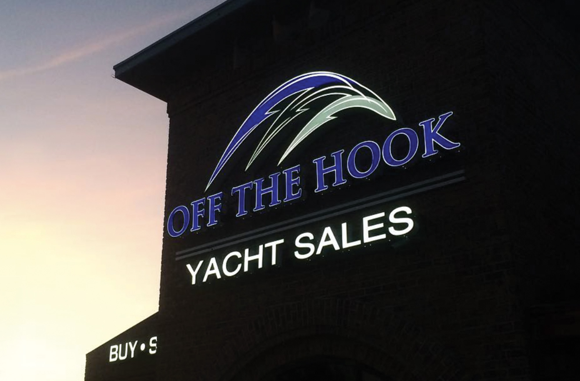 An exterior photograph of an illuminated sign for "OFF THE HOOK YACHT SALES" mounted on a brick building tower against a soft sunset sky.