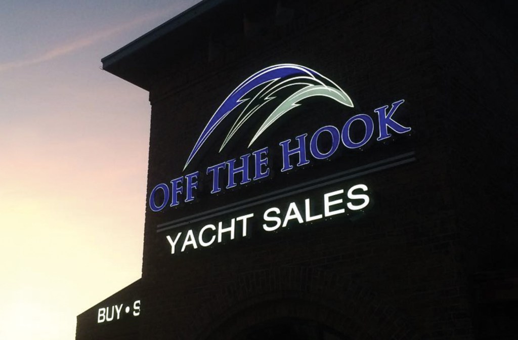 Screenshot An exterior photograph of an illuminated sign for "OFF THE HOOK YACHT SALES" mounted on a brick building tower against a soft sunset sky.