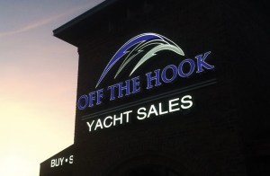 An exterior photograph of an illuminated sign for "OFF THE HOOK YACHT SALES" mounted on a brick building tower against a soft sunset sky.