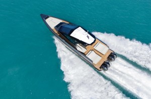 An aerial view of a sleek, metallic dark-gray Wallypower 50X powered by four Mercury outboard engines carving through bright turquoise ocean waters, showing off its spacious teak decks, large sunpads, and angular hardtop design.