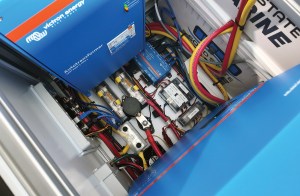 An overhead view into a compact marine electrical enclosure featuring Victron Energy components, including an autotransformer, circuit fuses, wiring harnesses, and marine battery terminals.