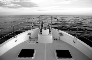 A black-and-white view looking out over the bow of a trawler yacht toward a calm, open ocean under a expansive sky at sea.