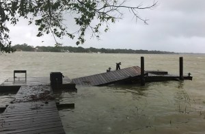A damaged wooden floating dock collapsed into rough, wind-rippled coastal waters under an overcast sky following a severe storm.