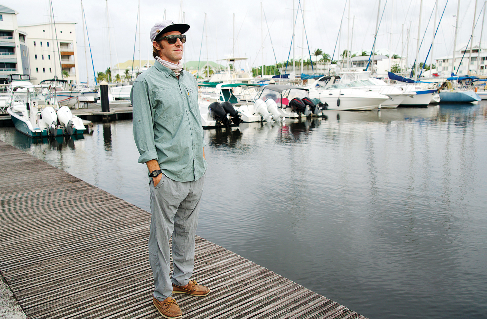 A man wearing a long-sleeve UPF shirt, flat-brim cap, sunglasses, and neck gaiter stands on a wooden dock surrounded by anchored boats in a marina.