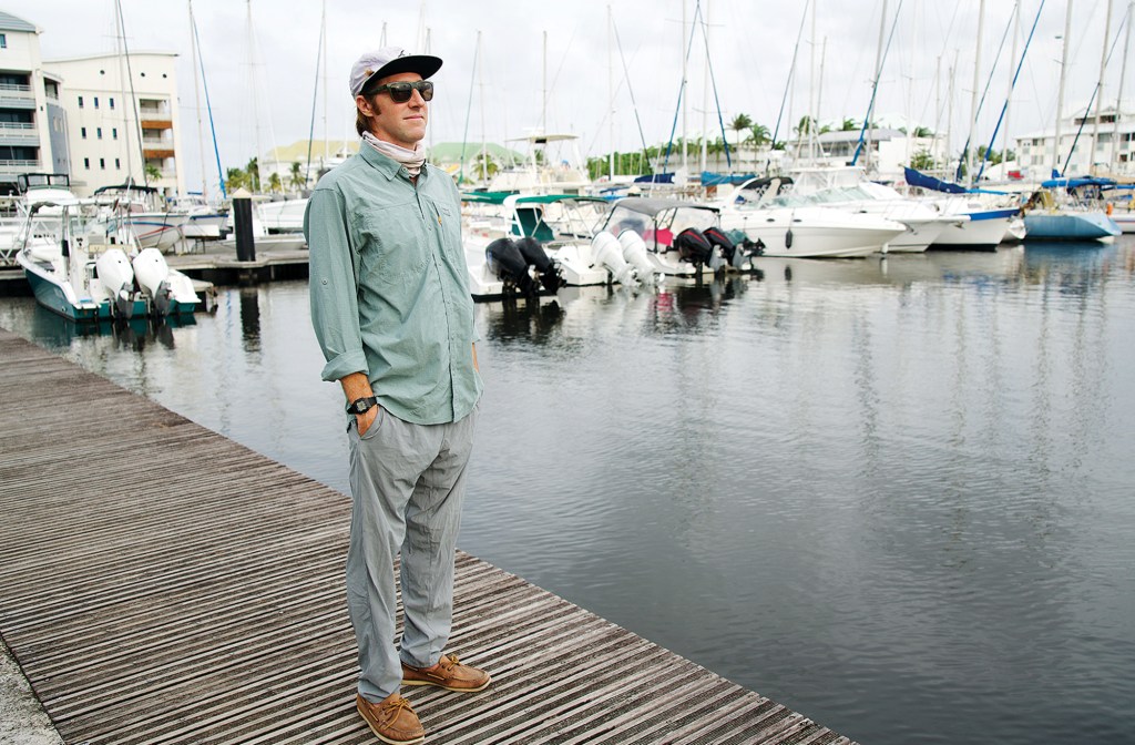 feat_Owen-fashionista A man wearing a long-sleeve UPF shirt, flat-brim cap, sunglasses, and neck gaiter stands on a wooden dock surrounded by anchored boats in a marina.