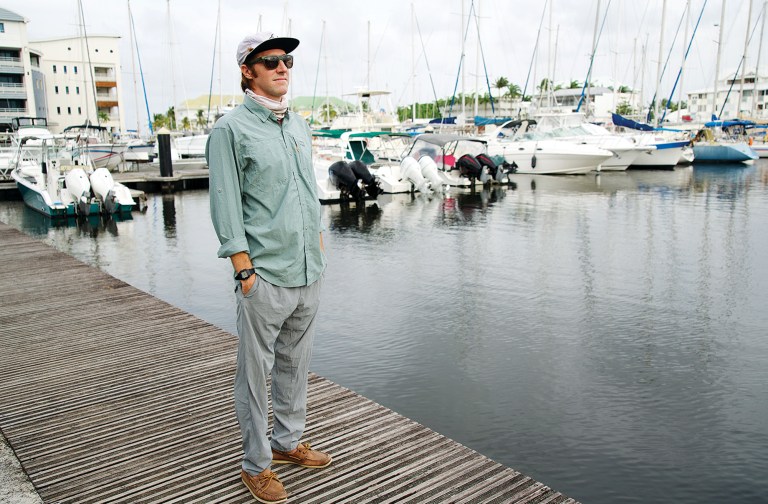 feat_Owen-fashionista A man wearing a long-sleeve UPF shirt, flat-brim cap, sunglasses, and neck gaiter stands on a wooden dock surrounded by anchored boats in a marina.