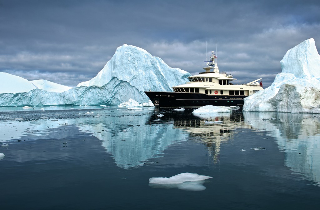 feat_Vripack-Beothuk The 102-foot explorer yacht Beothuk navigating calm, ice-filled Arctic waters surrounded by massive icebergs and dark overcast skies.