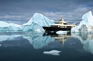 The 102-foot explorer yacht Beothuk navigating calm, ice-filled Arctic waters surrounded by massive icebergs and dark overcast skies.