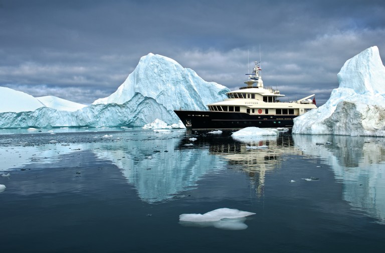 feat_Vripack-Beothuk The 102-foot explorer yacht Beothuk navigating calm, ice-filled Arctic waters surrounded by massive icebergs and dark overcast skies.