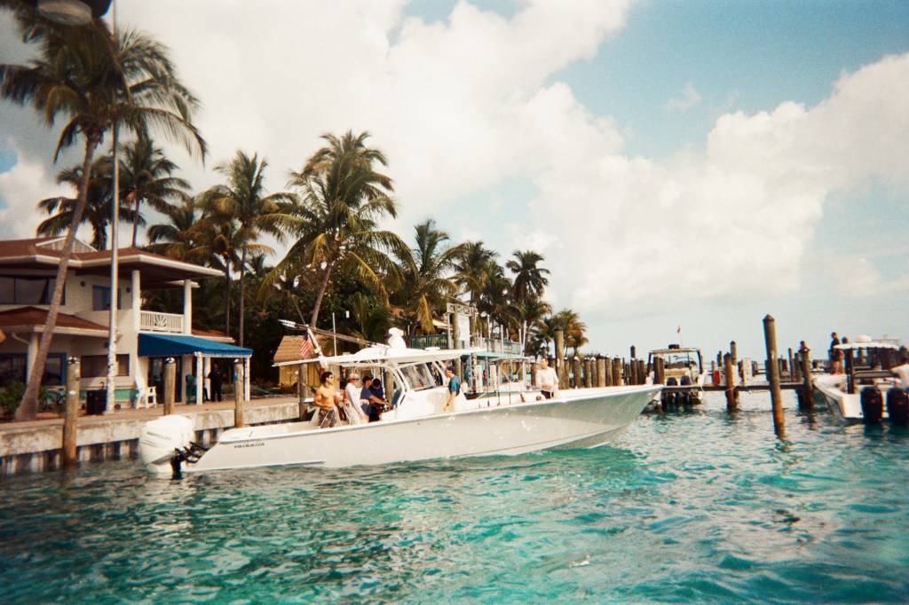 A white Valhalla center console powerboat carrying passengers near a wooden dock surrounded by palm trees and tropical waterfront buildings under a partly cloudy sky.