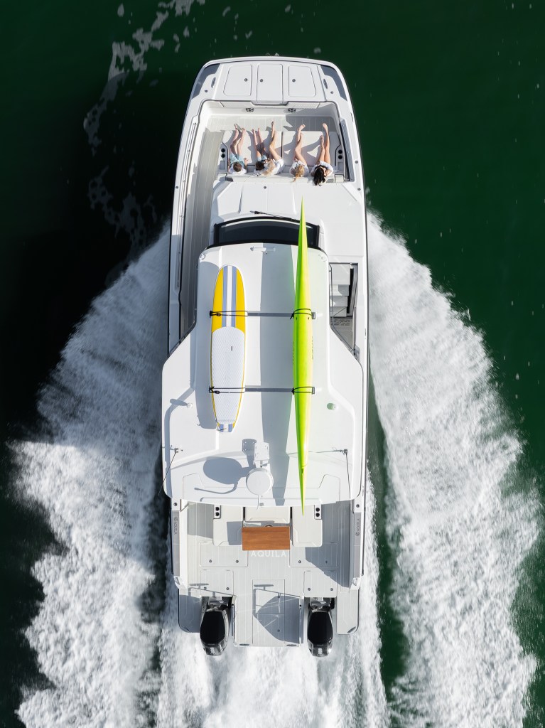 Direct top-down aerial view of an Aquila 45 Sport power catamaran cruising underway through open water, creating white wake trails along its twin hulls while passengers lounge on the bow sunpad and a surfboard and kayak are mounted to the hardtop roof rack.