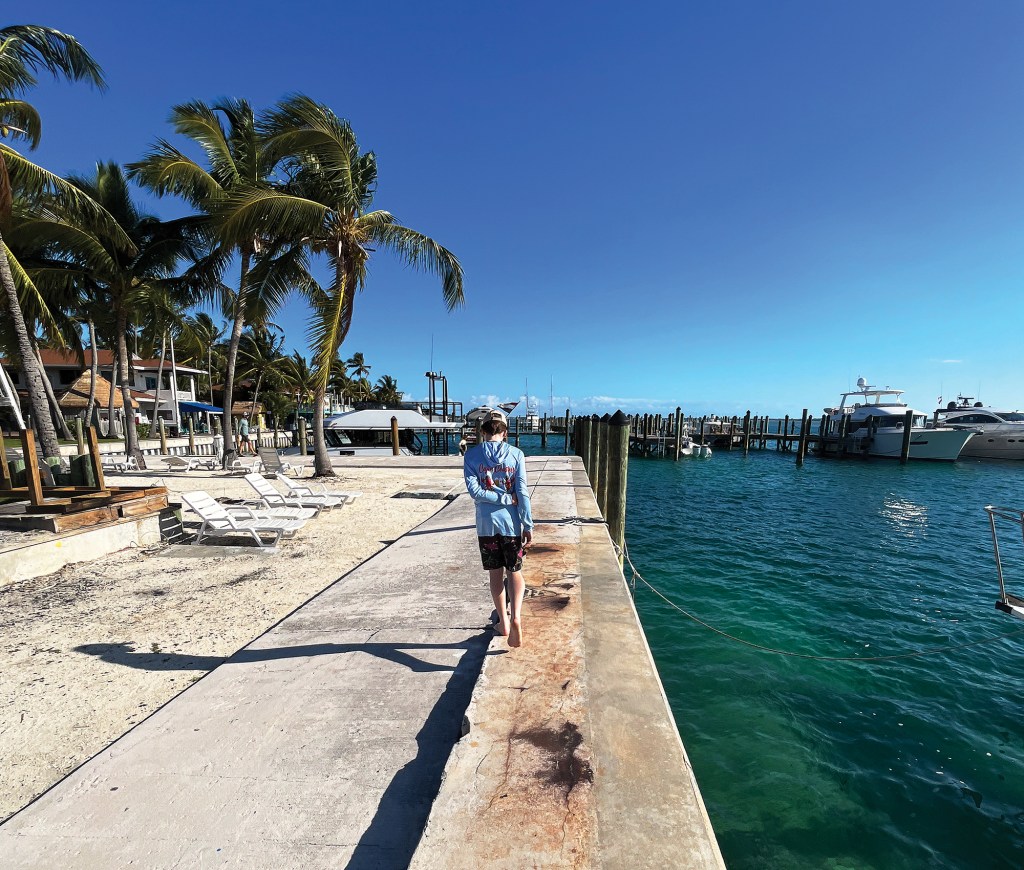 Person walking barefoot along a concrete dock lined with palm trees beside yachts at a tropical resort marina in The Bahamas.