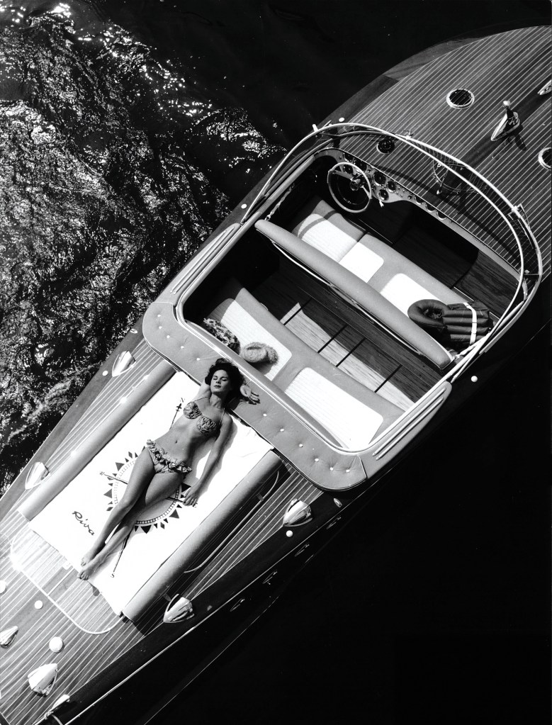 Vintage black-and-white aerial view of a woman sunbathing on the aft deck pad of a classic wooden Riva Aquarama.