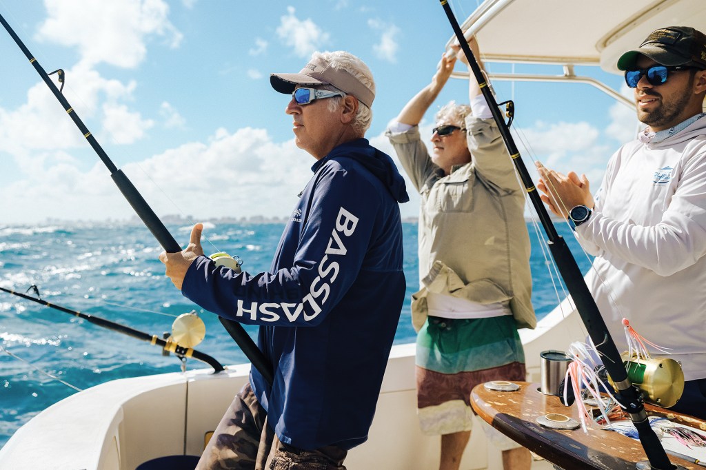 Three men deep-sea fishing aboard a boat on open ocean water, holding fishing rods and wearing sun-protective shirts, sunglasses, and hats.