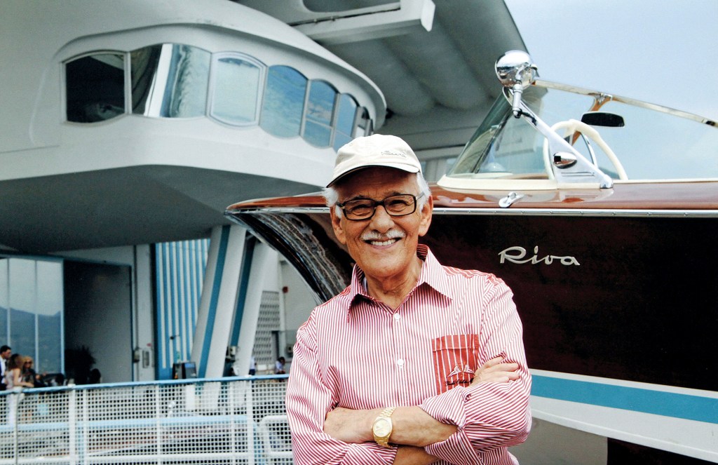 Carlo Riva smiling with arms crossed in front of a classic wooden Riva speedboat at the shipyard.